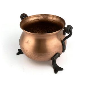 Puede incluir: Una pequeña olla de cobre con textura martillada y tres patas de hierro negro. La olla tiene dos asas de hierro negro.