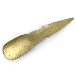 May include: A vintage brass shoe horn with a long, slender design. The shoe horn has a rounded scoop at one end and a pointed tip, with decorative detailing along the top edge. The brass has a warm, golden hue.