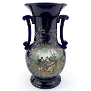 May include: A dark blue ceramic vase with two curved handles. The vase features a painted scene of people in a garden setting. The vase has a wide mouth and a rounded base, and is a decorative item.
