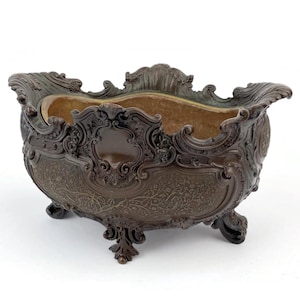 May include: An ornate bronze-coloured planter with a detailed floral and scroll design. The planter has a rectangular shape with curved sides, supported by three decorative feet. The interior has a light brown colour.