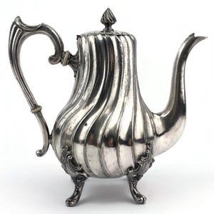 May include: A silver-colored teapot with a fluted body, curved handle, and a spout. The teapot has a decorative lid with a finial and rests on three ornate feet. The design is vintage and elegant, suitable for serving tea.