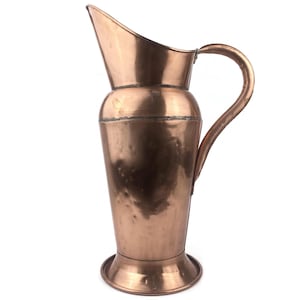 May include: A copper pitcher with a curved handle and a wide spout. The pitcher has a cylindrical body that tapers towards the base, which sits on a circular foot. The copper has a warm, reddish-brown hue, and the pitcher appears to be vintage.