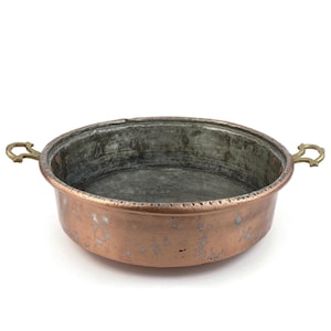 Op de afbeelding: Een koperen pan met messing handvatten. De pan heeft een ronde vorm met een licht verhoogde rand. Het koper heeft een warme, roodbruine kleur met enkele verkleuringen. De handvatten zijn glanzend goudkleurig.