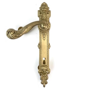 Pode incluir: Puxador de porta de latão dourado ornamentado com um design decorativo. A maçaneta apresenta uma alavanca curva e uma placa traseira longa e retangular com detalhes intrincados, incluindo motivos florais e de rolagem. O buraco da fechadura é visível.