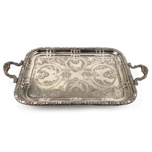 May include: A rectangular silver-toned serving tray featuring ornate, embossed floral and scrollwork patterns across its surface. The tray includes two decorative, curved handles on opposite sides and a raised, scalloped edge with detailed metalwork.