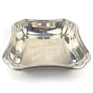 Könnte beinhalten: Eine quadratische, silberfarbene Servierschale mit gewelltem Rand. Die Schale hat eine reflektierende Oberfläche und eine leicht gebogene Form. Das Design ist schlicht und elegant, geeignet zum Servieren von Speisen oder als dekoratives Stück.