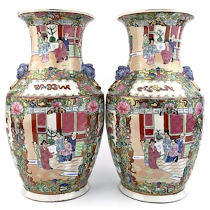 Puede incluir: Par de jarrones de porcelana ornamentados, pintados a mano. Cada jarrón presenta una escena detallada con figuras vestidas con trajes tradicionales, rodeadas de patrones florales y geométricos. Los jarrones tienen un borde acampanado y asas con forma de cabeza de león, con una paleta de colores rosa, verde y dorado.