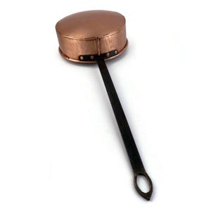 Op de afbeelding: Een gehamerde koperen lepel met een lange, zwarte ijzeren steel. De lepel heeft een ronde kom en een kleine lus aan het einde van de steel.