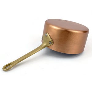 Könnte beinhalten: Ein Kupferkochtopf mit einem Messinggriff. Der Topf hat einen abgerundeten Körper und einen langen Griff, der mit Nieten befestigt ist. Die Kupferoberfläche reflektiert das Licht, und der Griff hat eine Schlaufe zum Aufhängen. Ein klassischer Küchenartikel.