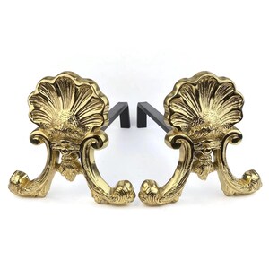 May include: Pair of ornate brass fireplace andirons. Each andiron features a shell-like design with a black support bar. The andirons have a curved base and are made of a shiny, gold-colored metal.