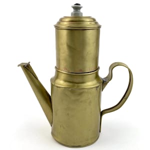 May include: A vintage brass coffee pot with a spout, handle, and lid. The pot has a cylindrical shape with a wider base and a narrower top section. The lid has a small, gray knob. The coffee pot is made of brass and has a shiny, golden finish.