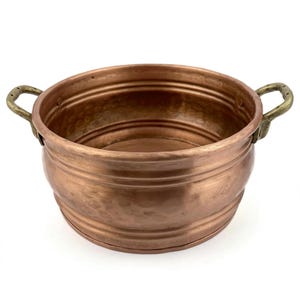 May include: A copper-colored bowl with two brass handles. The bowl has a rounded shape with a slightly flared rim and horizontal ridges around the body. The handles are curved and attached to the sides of the bowl.