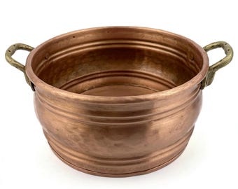 May include: A copper bowl with two brass handles. The bowl has a rounded shape with a slightly flared rim and horizontal ridges around the body. The handles are curved and attached to the sides of the bowl.