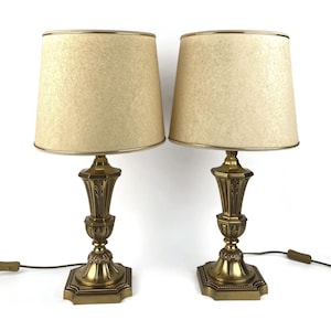 Może przedstawiać: Dwie pasujące lampy stołowe w stylu vintage. Każda lampa ma beżowy abażur ze złotym wykończeniem. Podstawa wykonana jest ze szczegółowego metalu w kolorze złotym z kwadratową podstawą. Lampy są podłączone do gniazdka.