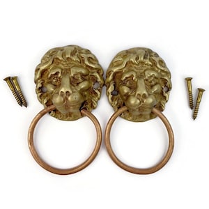 May include: Two antique brass lion head door knockers with copper-colored rings. The lion heads are detailed with textured manes and facial features. Includes brass screws for installation. Decorative hardware.