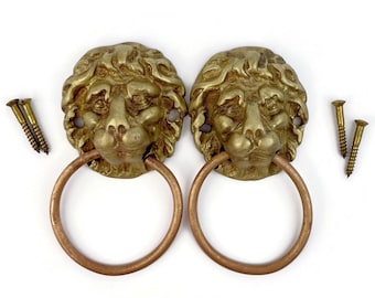 Puede incluir: Dos aldabas antiguas de latón con forma de cabeza de león y anillos cobrizos. Las cabezas de león tienen melenas y rasgos faciales detallados. Incluye tornillos de latón. Herrajes decorativos.