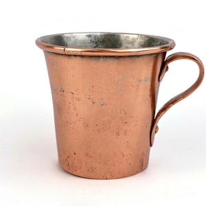 Puede incluir: Una taza de cobre con asa. La taza tiene una forma ligeramente cónica y un acabado metálico brillante. El interior de la taza tiene un forro plateado. El asa es curva y está unida al lateral de la taza. La taza probablemente se usa para beber.