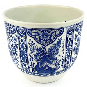 Puede incluir: Una maceta de cerámica con base blanca y complejos patrones florales azules dispuestos en paneles verticales. El diseño incluye flores estilizadas, hojas y motivos de volutas. El recipiente tiene una abertura ancha y una base acanalada.