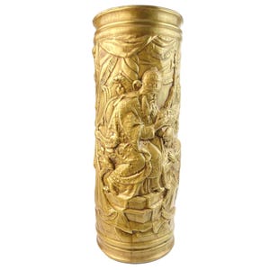May include: A gold-colored, cylindrical vase with detailed relief carvings. The carvings depict figures in ornate robes and headwear, possibly religious or historical figures. The vase has a textured surface and a slightly flared top and base.