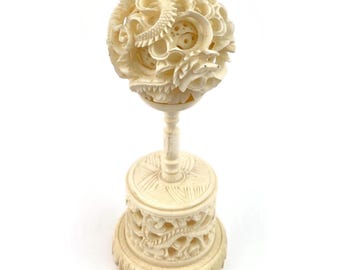 May include: Ivory-colored, intricately carved sphere sculpture with a dragon design. The sphere sits atop a pedestal with a detailed base. The sculpture is a decorative piece.