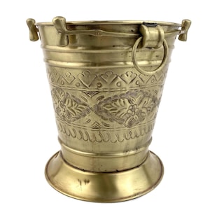 Puede incluir: Un cubo de metal dorado con un diseño floral en relieve. El cubo tiene un asa con un anillo en el lateral.