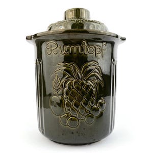 Könnte beinhalten: Dunkelgrüner Keramik-Rumtopf mit Deckel. Der Topf hat geprägte Ananas- und Fruchtdesigns. Das Wort "Rumtopf" steht auf der Vorderseite. Der Topf hat einen Griff auf jeder Seite und einen dekorativen Deckel.