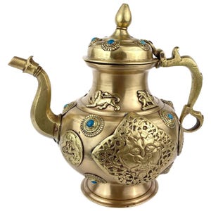Puede incluir: Una tetera de latón con intrincados detalles en relieve y detalles turquesas. La tetera presenta un pico curvo, un asa y una tapa con un remate. Los elementos decorativos incluyen figuras de leones e incrustaciones circulares turquesas. El diseño es ornamentado y detallado.