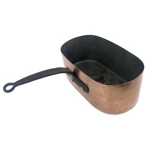 May include: Copper saucepan with a long, black handle. The pan has a rectangular shape with rounded corners and a dark interior. The handle is attached with a small, dark metal bracket. The pan is made of copper and has a shiny, reflective surface.