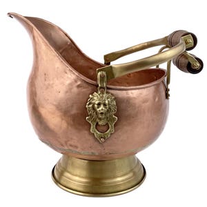 May include: A copper coal scuttle with a brass base and handle. The scuttle features a lion's head emblem on the front. The handle has wooden grips and brass accents. The scuttle is designed with a spout for easy pouring.