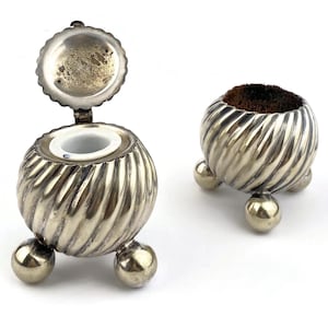 May include: Two antique silver-toned items. One is a small container with a hinged lid, the other is a pincushion. Both have a ribbed design and sit on three ball feet. The container's lid is open, revealing a white interior.