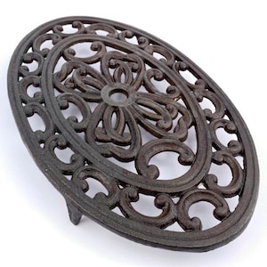 Puede incluir: Salvamanteles ovalado de hierro fundido marrón oscuro con un diseño floral y de volutas ornamentado. El salvamanteles tiene un motivo floral central y un borde decorativo. Cuenta con un pequeño pie para elevarlo y está diseñado para proteger las superficies del calor.