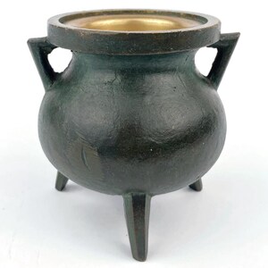 May include: A small, dark green metal cauldron with three legs and two handles. The interior of the cauldron is gold-colored. The cauldron has a rounded body and a wide rim.