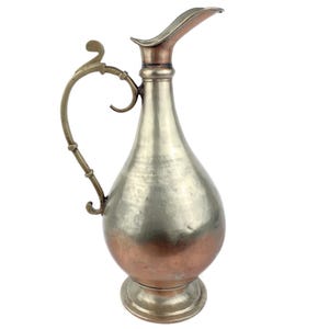 May include: A vintage metal jug with a silver body and a copper-coloured base. The handle is a decorative brass colour, and the spout has a copper finish. The jug has a bulbous shape and a wide base.