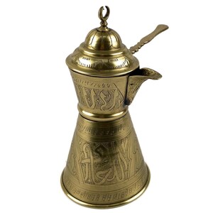 Op de afbeelding: Een koperen koffiepot met een conische voet en een rond deksel bekroond met een halve maan. De buitenkant is versierd met gegraveerde Arabische kalligrafie en geometrische patronen, inclusief een handvat aan de zijkant en een kleine tuit.