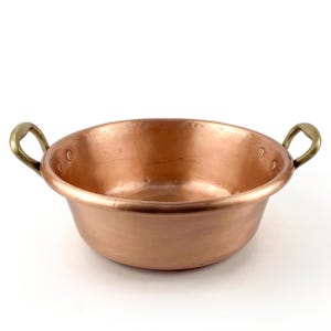 Peut inclure: Une bassine ronde en cuivre poli avec une finition métallique lisse et une large ouverture. Le récipient est doté de deux poignées incurvées en laiton, chacune fixée solidement aux parois par deux rivets visibles sur le bord intérieur.