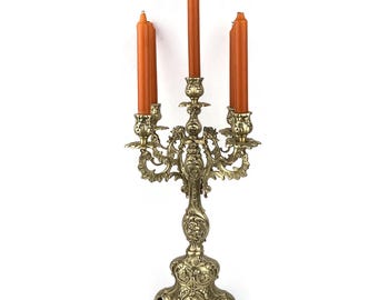 May include: A gold metal candelabra with five candle holders and ornate details. The candelabra is holding five orange candles.