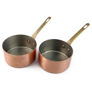 May include: Two round copper saucepans with silver-toned interior linings and long, angled brass handles. Each handle is securely attached to the side of the pan with two visible metal rivets. The exterior of the pans features a warm copper finish.