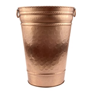 May include: A copper-colored metal bucket with a hammered texture. The bucket has a cylindrical shape with a slightly flared top and a decorative rope-like band. Two small ring handles are attached near the top.