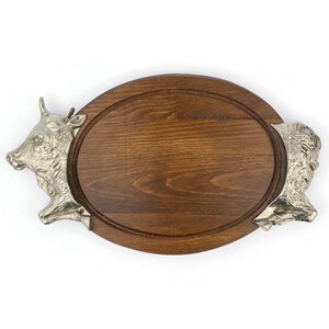 Op de afbeelding: Ovale houten serveerplank met een donkerbruine afwerking. De plank heeft zilverkleurige metalen accenten in de vorm van stieren- en bizonskoppen aan elk uiteinde. Er loopt een groef rond de rand van de plank.
