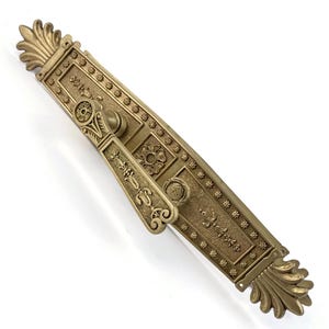 May include: Ornate antique brass door handle with intricate floral and scrollwork details. The rectangular backplate features a central floral motif and is flanked by rows of small flower-shaped embellishments. The handle itself has a decorative lever.
