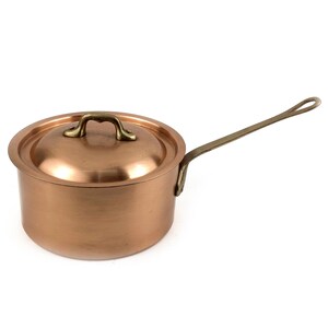 Puede incluir: Una cacerola de cobre con tapa y mango largo. La olla tiene una superficie brillante y un mango de color latón. La tapa tiene un asa de latón. La cacerola está diseñada para cocinar y probablemente se usa en una cocina.