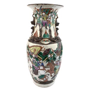 Puede incluir: Un jarrón alto de cerámica blanquecina con borde acampanado y base ancha. El jarrón presenta una escena pintada detallada y colorida de figuras con vestimenta tradicional. Los detalles y asas de color marrón oscuro añaden contraste. El jarrón es decorativo.