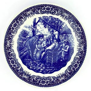 May include: A round, white and blue decorative plate. The plate features a detailed blue illustration of a gathering of people, with a floral border. The illustration depicts a group of people gathered around a barrel, with a horse nearby.