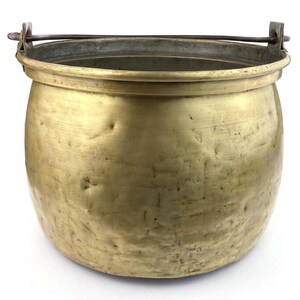 Puede incluir: Un caldero de latón vintage con un cuerpo redondeado y un asa resistente. El caldero tiene un aspecto deslustrado y envejecido, con abolladuras e imperfecciones visibles. El asa es de metal oscuro, y el diseño general sugiere una estética rústica o antigua.