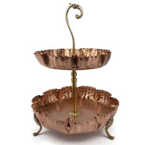 Könnte beinhalten: Zweistöckiger Servierständer aus Kupfer mit Messinggriff und Akzenten. Die Kupferschalen haben einen gerüschten Rand und eine gehämmerten Textur. Der Ständer hat dekorative Messingfüße und eine zentrale Stütze.