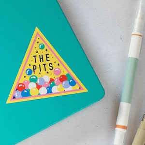 May include: A teal notebook with a yellow triangle sticker that says "THE PITS" with colorful dots inside. Two pens, one white and one brown, are next to the notebook on a white surface.