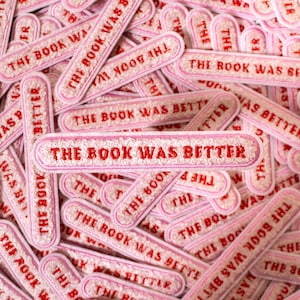 以下が含まれることがあります： 「The Book Was Better」と書かれたピンクと赤の刺繍入りアイロン接着パッチの山をクローズアップした写真です。