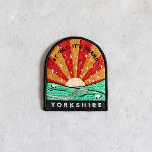 May include: A black and yellow embroidered patch with a red and yellow sun rising over a green field with white sheep. The text "By Heck It's Grand Yorkshire" is written around the sun.