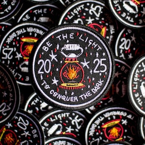 May include: A black and white embroidered patch with the text "Be the light to conquer the dark" and a red and orange lantern with flames. The patch has white stars and the numbers 20 and 25.