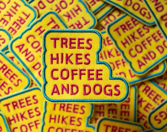 May include: A close-up of several yellow fabric patches with blue stitching. The patches have the text "Trees, Hikes, Coffee and Dogs" in pink and blue lettering.